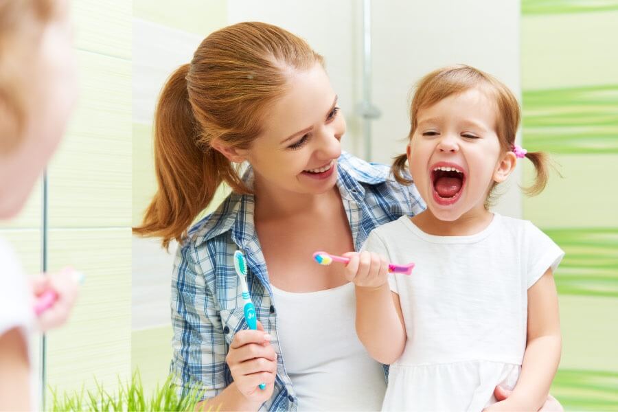 Blonde mom teaches her young daughter how to brush and floss her teeth in a green and white bathroom